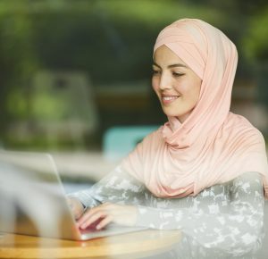 Pretty positive young woman in pink hijab answering e-mails on laptop